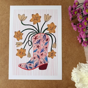 May include: A pink cowboy boot with a floral pattern is filled with yellow flowers. The boot is on a pink and white striped background.