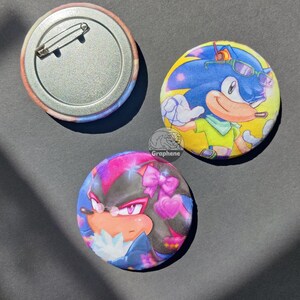 May include: Three circular pin-back buttons. One shows the back with a metal pin. Another features a blue hedgehog character with green shirt and sunglasses. The third displays a black hedgehog character with pink bows and hearts. Text reads Graphene.