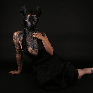 Puede incluir: Una persona con una m&aacute;scara de perro de cuero negro con tachuelas plateadas y un collar negro con una cadena plateada. Lleva un vestido negro y tiene tatuajes en los brazos.