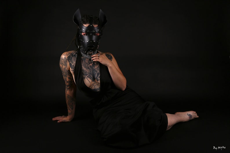 Puede incluir: Una persona con una m&aacute;scara de perro de cuero negro con tachuelas plateadas y un collar negro con una cadena plateada. Lleva un vestido negro y tiene tatuajes en los brazos.