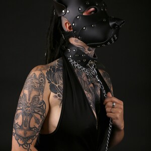 Puede incluir: Una persona lleva una m&aacute;scara de perro de cuero negro con tachuelas plateadas y un collar de cuero negro con una cadena plateada. La persona tiene tatuajes en los brazos y el pecho.
