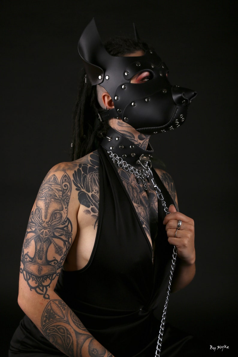 Puede incluir: Una persona lleva una m&aacute;scara de perro de cuero negro con tachuelas plateadas y un collar de cuero negro con una cadena plateada. La persona tiene tatuajes en los brazos y el pecho.