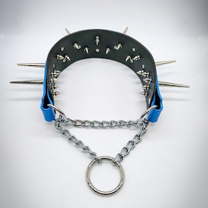 May include: Black leather collar with silver spikes and studs, blue accents, and a silver chain with a large ring. The collar has long, sharp spikes extending outwards. The design is edgy and bold.
