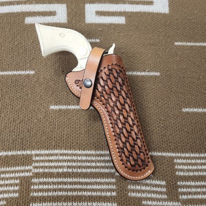May include: A vintage-style revolver with a cream-colored handle is secured in a brown leather holster. The holster features a woven design and a snap closure. The overall aesthetic suggests a Western or historical theme.