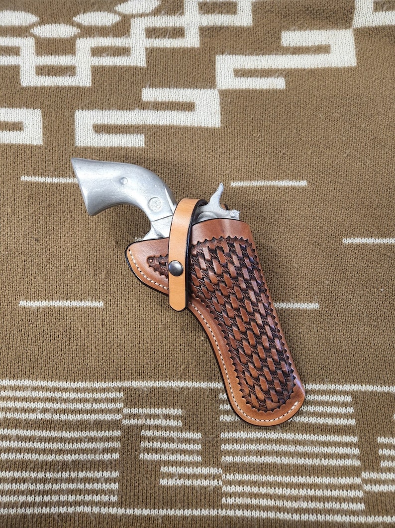 May include: A brown leather holster with a woven pattern for a silver revolver. The holster has a strap with a metal snap.