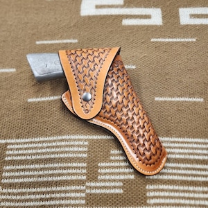 May include: A light brown leather holster with a woven basket weave pattern. The holster has a snap closure and holds a silver-colored object. The background is a brown textile.