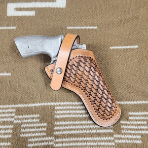 May include: A silver handgun in a light brown leather holster with a basket weave pattern. The holster has a strap with a snap closure. The background is a brown and white patterned fabric.