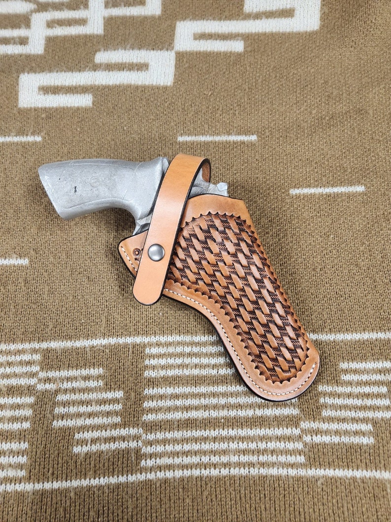 May include: A silver handgun in a light brown leather holster with a basket weave pattern. The holster has a strap with a snap closure. The background is a brown and white patterned fabric.