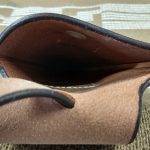 May include: A brown leather pouch with a dark brown trim and a snap closure. The interior is a lighter brown suede-like material. The pouch has a loop on the side. The background is a patterned fabric.