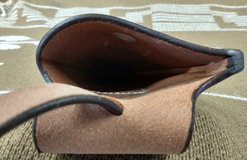 May include: A brown leather pouch with a dark brown trim and a snap closure. The interior is a lighter brown suede-like material. The pouch has a loop on the side. The background is a patterned fabric.