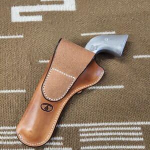 May include: A brown leather holster with a white stitching detail and a logo of a tree. The holster is designed to hold a silver revolver.