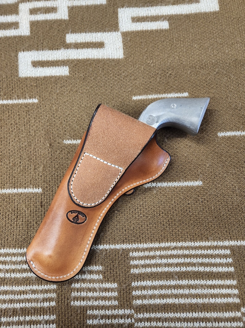 May include: A brown leather holster with a white stitching detail and a logo of a tree. The holster is designed to hold a silver revolver.