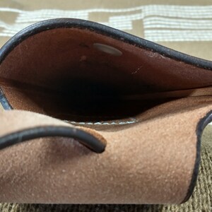 May include: A brown leather pouch with a black leather strap. The pouch is open and the inside is visible. The pouch is made of a soft, supple leather.
