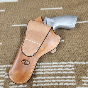 May include: A light brown leather holster with white stitching and a silver-colored handgun. The holster has a stamped logo and is designed to hold the firearm securely. The background is a tan and white patterned fabric.