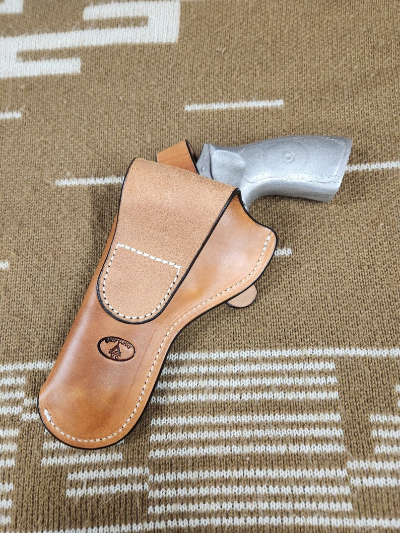 May include: A light brown leather holster with white stitching and a silver-colored handgun. The holster has a stamped logo and is designed to hold the firearm securely. The background is a tan and white patterned fabric.