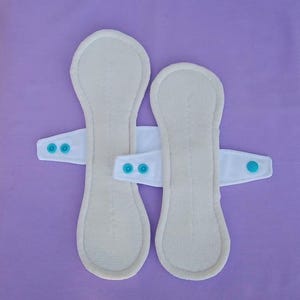 Può includere: Due assorbenti mestruali riutilizzabili in tessuto bianco sporco dalla forma curva e allungata. Ogni assorbente ha una linguetta bianca con due bottoni a pressione turchese per il fissaggio. Su sfondo lilla chiaro.