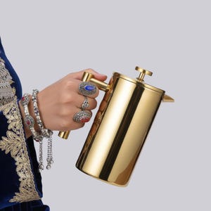 May include: A polished gold-tone stainless steel French press coffee maker with a cylindrical body, a flat lid, and a plunger mechanism. The metallic finish reflects light, highlighting the smooth, vertical surface of the carafe and handle.