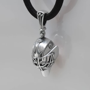 Peut inclure: Un collier avec un pendentif en argent représentant un masque de gardien de hockey détaillé. Le masque est en métal et est sur un cordon noir.