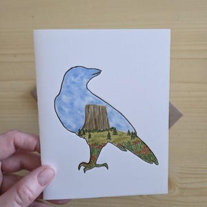 May include: A watercolor greeting card featuring a blue bird silhouette with a landscape scene inside. The landscape includes a brown rock formation, green trees, and red and orange flowers. The card is held by a hand.