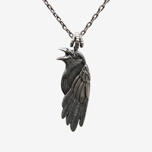 May include: A silver chain necklace with a detailed, three-dimensional crow pendant. The crow is depicted with its wings spread and its beak open.