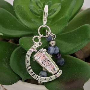 Op de afbeelding: Zilveren bedel met een koffiekopontwerp en het woord "COFFEE" eromheen. De bedel is bevestigd aan een zilveren ketting met blauwe en paarse kralen.