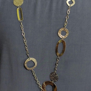 Op de afbeelding: Een lange zilverkleurige ketting met een reeks afwisselende geometrische vormen, waaronder gehamerde cirkels, gladde ovalen en vierkanten verbonden door een fijne ketting, eindigend in een doorzichtige, ronde vergrootglas hanger.