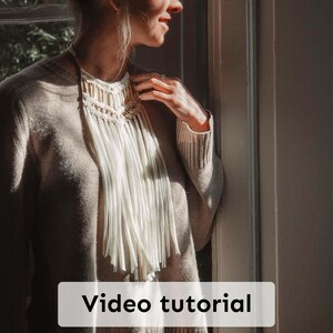 May include: A cream-coloured macrame necklace with a fringe design. The necklace has a woven top section with gold-coloured accents and long, flowing fringe. The text "Video tutorial" is displayed on the bottom of the image.