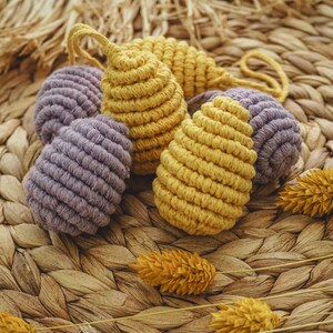 May include: A group of five handmade macrame ornaments in a light purple and yellow color. The ornaments are made with a thick cord and have a textured, woven appearance. The ornaments are displayed on a woven wicker surface.