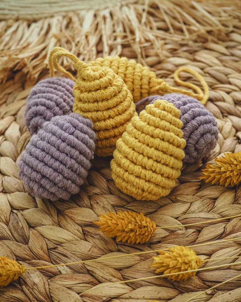 May include: A group of five handmade macrame ornaments in a light purple and yellow color. The ornaments are made with a thick cord and have a textured, woven appearance. The ornaments are displayed on a woven wicker surface.