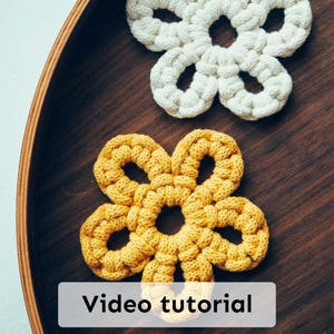 May include: Two handmade, flower-shaped crochet coasters. One is cream-colored, the other is yellow. The coasters are placed on a dark wooden tray. The text "Video tutorial" is displayed on a white rectangle.