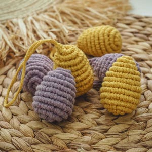 May include: A group of five crocheted eggs in a woven basket. Two of the eggs are a light purple color and three are a mustard yellow color.