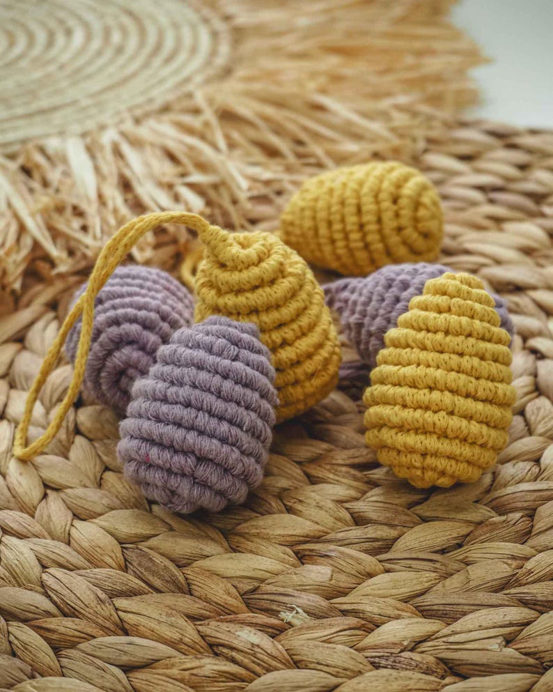 May include: A group of five crocheted eggs in a woven basket. Two of the eggs are a light purple color and three are a mustard yellow color.