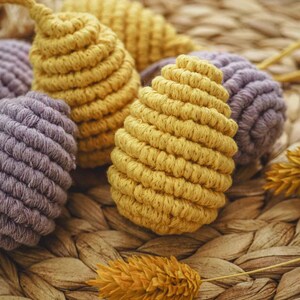 May include: Close-up of a group of handmade macrame ornaments in yellow and purple. The ornaments are made with a thick, textured cord and have a woven, rope-like appearance. The ornaments are displayed on a woven wicker surface.