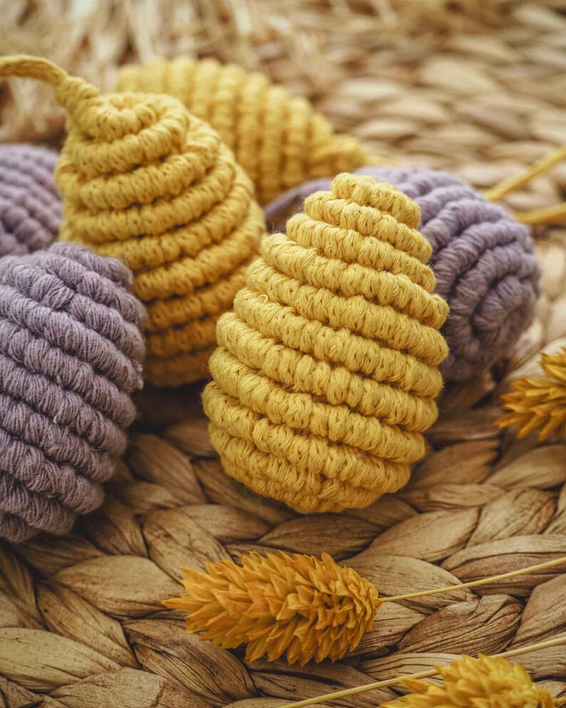 May include: Close-up of a group of handmade macrame ornaments in yellow and purple. The ornaments are made with a thick, textured cord and have a woven, rope-like appearance. The ornaments are displayed on a woven wicker surface.