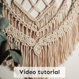 以下が含まれることがあります： クリーム色のマクラメの壁掛け。複雑な結び目の模様とフリンジが特徴です。「Video tutorial」というテキストが表示されています。木製のダボに取り付けられています。装飾用のストリングライトが上に吊るされています。
