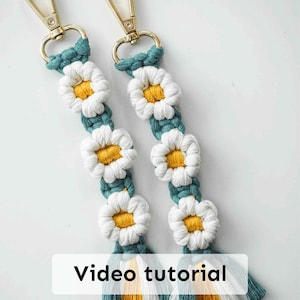 以下が含まれることがあります： デイジーの花のデザインが施された、2つのマクラメキーホルダー。各キーホルダーは、黄色の中心部を持つ白い花、ターコイズのアクセント、金色の留め金が特徴です。各キーホルダーの下部には、ターコイズ、黄色、白の糸で作られたタッセルが付いています。「Video tutorial」というテキストが画像にあります。