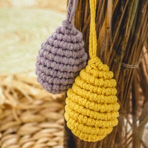 May include: Two crocheted egg-shaped ornaments hanging from a wooden branch. One ornament is purple and the other is yellow.