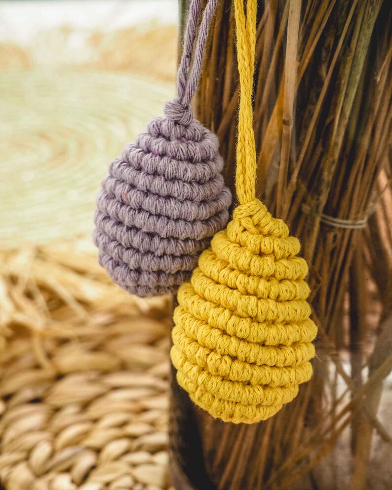 May include: Two crocheted egg-shaped ornaments hanging from a wooden branch. One ornament is purple and the other is yellow.