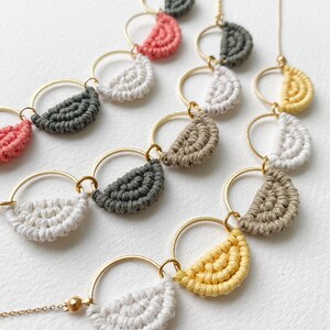 May include: A gold chain necklace with multiple layered circles, each featuring a half-circle of macrame in different colours, including white, grey, yellow, and pink.