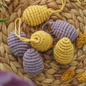May include: Six crocheted Easter eggs in yellow and purple. The eggs are made with a thick yarn and have a textured surface. They are arranged on a woven wicker surface.