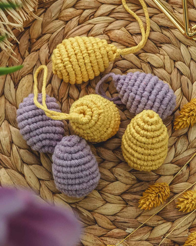 May include: Six crocheted Easter eggs in yellow and purple. The eggs are made with a thick yarn and have a textured surface. They are arranged on a woven wicker surface.