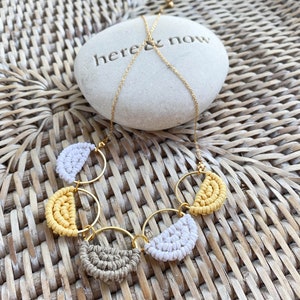 May include: A gold chain necklace with three macrame circles, each with a half-circle of yellow, white and brown macrame. The necklace is on a woven wicker surface with a white stone that says "here & now".