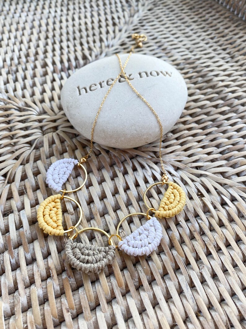 May include: A gold chain necklace with three macrame circles, each with a half-circle of yellow, white and brown macrame. The necklace is on a woven wicker surface with a white stone that says "here & now".