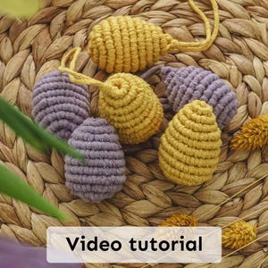 May include: A collection of six macrame Easter eggs, four yellow and two purple, with a woven texture. The eggs are hanging from a woven straw mat with the text "Video tutorial" on a white rectangle.