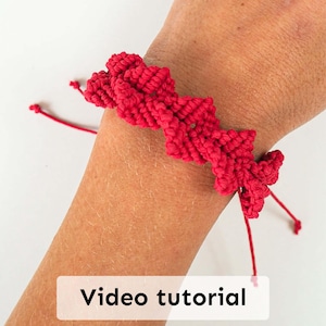 以下が含まれることがあります： 手作りの赤いマクラメブレスレット。ジグザグ模様で、手首に装着されています。結び目の糸で作られ、調節可能な紐が付いています。「Video tutorial」の文字が画像に表示されています。