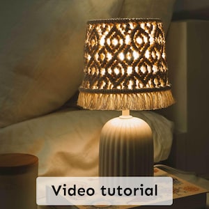 Può includere: Una lampada in ceramica bianca con un paralume in macramé intrecciato con un bordo a frangia. La lampada è su un comodino con un libro e una candela.