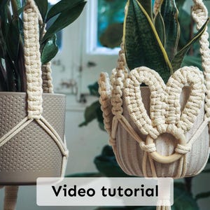 May include: Two beige macrame plant hangers with potted plants. The hangers have a woven design and wooden rings. The pots are a light gray color. The text "Video tutorial" is displayed on a white rectangle at the bottom of the image.
