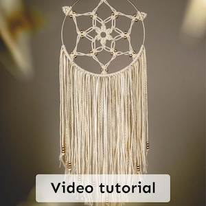 May include: Cream-coloured macrame dream catcher with a circular gold metal frame. The dream catcher features a central starburst design, gold beads, and long, flowing fringe. The text "Video tutorial" is displayed on a white rectangle at the bottom.