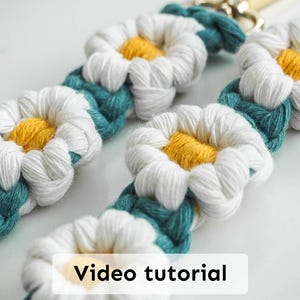 May include: A white and teal macrame keychain with yellow daisy flowers. The keychain has a gold metal clasp. "Video tutorial" is written in white text on a black background.