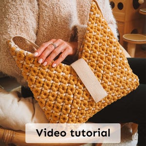 May include: A handmade, mustard yellow crochet bag with white accents and a beige strap. The bag features a unique, textured pattern and a rectangular beige strap closure. The words "Video tutorial" are displayed on the bag.
